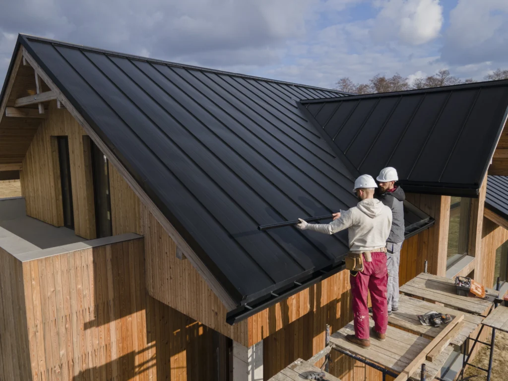 Roof replacement in progress as two workers install metal roofing panels on a modern wooden house while standing on scaffolding.