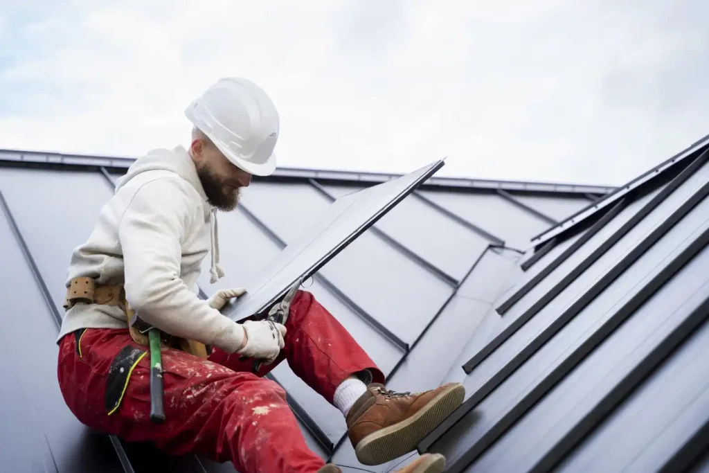 Worker in a white hard hat inspecting a metal roof panel for leaks while seated on a sloped rooftop.
