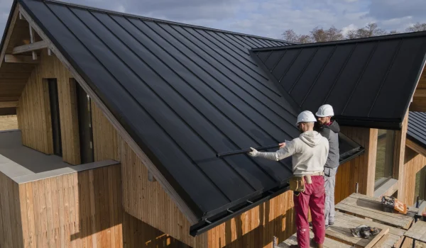 Roof replacement in progress as two workers install metal roofing panels on a modern wooden house while standing on scaffolding.