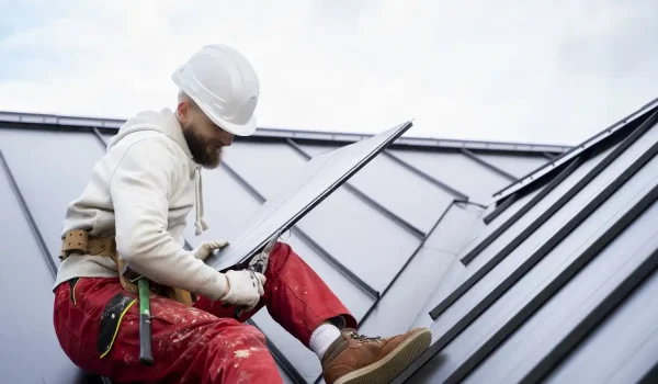 Worker in a white hard hat inspecting a metal roof panel for leaks while seated on a sloped rooftop.